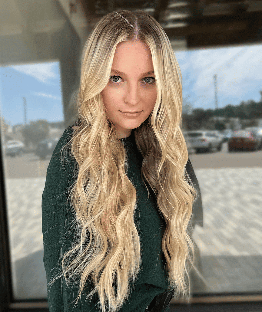 A person with long, wavy blonde hair wearing a dark green top stands near a window.