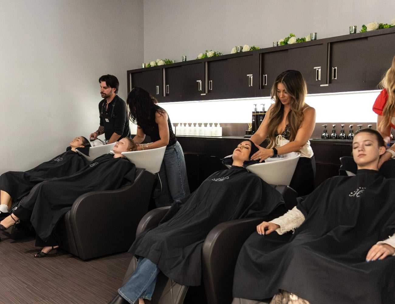 Hair salon with stylists washing clients' hair in modern black chairs.
