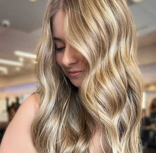 Young woman with long, wavy blonde hair in a bright salon setting.